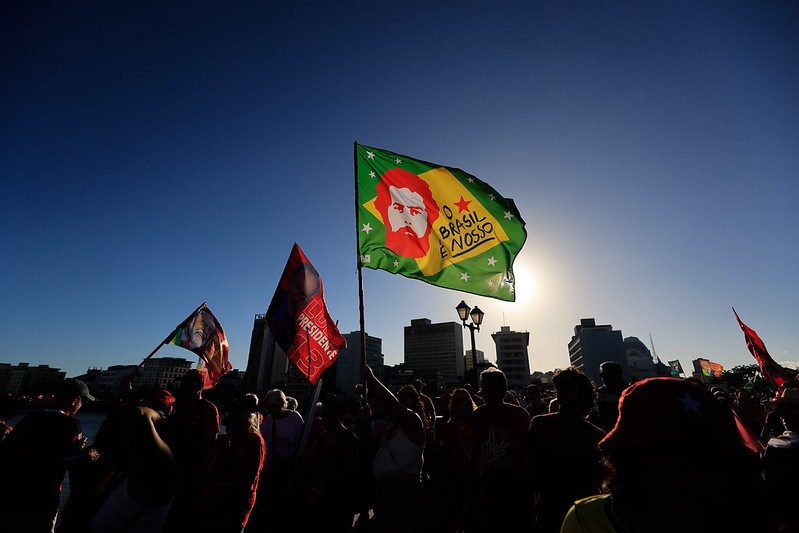 Fotografia de uma manifestação ao ar livre no final da tarde, com várias pessoas reunidas em primeiro plano, vistas principalmente em silhueta contra a luz do sol. Ao centro, destaca-se uma grande bandeira verde com a frase “Brasil é nosso” e a ilustração estilizada de uma figura humana, enquanto outras bandeiras vermelhas são erguidas ao redor. Ao fundo, aparecem edifícios urbanos e postes de iluminação sob um céu azul limpo, criando forte contraste entre a luminosidade do horizonte e as pessoas na cena.