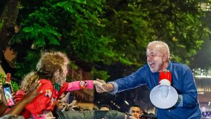 Foto noturna de uma multidão reunida ao ar livre em que o presidente Lula, de camisa azul, segurando um megafone vermelho e branco, com um largo sorriso no rosto inclina-se sobre uma barreira para cumprimentar pessoas que estendem as mãos em sua direção, com destaque para uma menina, de cabelos castanhos. Ao fundo, há árvores iluminadas por luzes verdes e diversos espectadores observando a cena, criando um ambiente movimentado e de interação próxima entre Lula e o público.