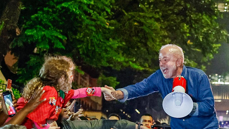Foto noturna de uma multidão reunida ao ar livre em que o presidente Lula, de camisa azul, segurando um megafone vermelho e branco, com um largo sorriso no rosto inclina-se sobre uma barreira para cumprimentar pessoas que estendem as mãos em sua direção, com destaque para uma menina, de cabelos castanhos. Ao fundo, há árvores iluminadas por luzes verdes e diversos espectadores observando a cena, criando um ambiente movimentado e de interação próxima entre Lula e o público.