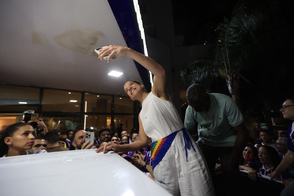 Fotografia noturna mostrando Raquel Lyra em destaque, vestida com roupa branca e uma faixa colorida na cintura, inclinando-se sobre um veículo branco enquanto ergue um celular para tirar uma selfie. Ao redor, há uma multidão reunida, muitas pessoas também segurando celulares para registrar o momento. A cena acontece em frente a um edifício iluminado, com palmeiras visíveis ao fundo e iluminação artificial destacando os participantes e a atmosfera de um evento público.