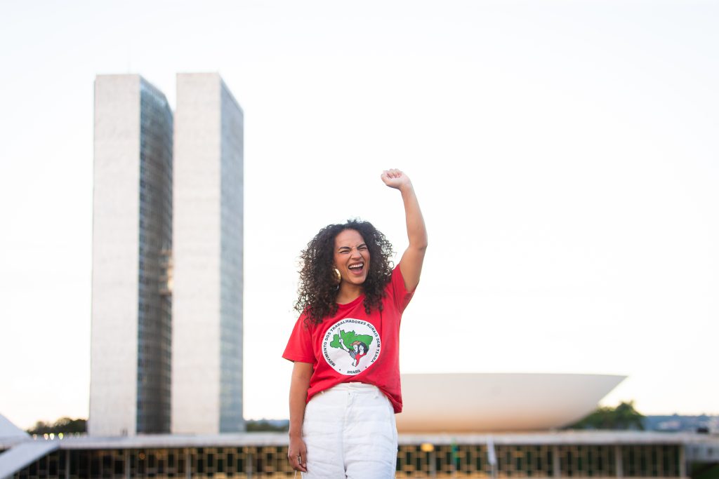 Rosa Amorim, uma mulher sorridente de longos cabelos encaracolados, vestindo uma camiseta vermelha do Movimento dos Trabalhadores Rurais Sem Terra (MST) e calça branca, levanta o braço em sinal de comemoração em frente ao Congresso Nacional, em Brasília.