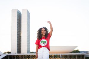 Rosa Amorim, uma mulher sorridente de longos cabelos encaracolados, vestindo uma camiseta vermelha do Movimento dos Trabalhadores Rurais Sem Terra (MST) e calça branca, levanta o braço em sinal de comemoração em frente ao Congresso Nacional, em Brasília.