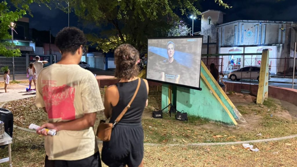 Foto noturna em uma praça ou espaço público, com duas pessoas observando uma tela de projeção montada ao ar livre ao lado de um brinquedo de playground. A tela exibe o rosto de uma pessoa durante uma apresentação em vídeo, enquanto, ao redor, há árvores, postes de iluminação, algumas pessoas circulando e prédios ao fundo, criando um cenário de exibição audiovisual comunitária em ambiente aberto.