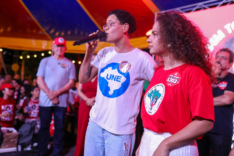 Foto de um evento público realizado sob uma tenda colorida, com iluminação em tons de vermelho e azul. Em primeiro plano, um jovem, magro, de cabelos crespos escuros, usando camiseta branca com o logotipo “UNE” fala ao microfone, enquanto Rosa Amorim, mulher negra de cabelos castanhos encaracolados, usando camiseta vermelha com adesivos e um emblema estampado, está ao lado. Ao fundo, há várias pessoas observando a cena, algumas também vestidas de vermelho. O ambiente parece ser uma atividade coletiva ou encontro com público, com foco na fala da pessoa que segura o microfone.