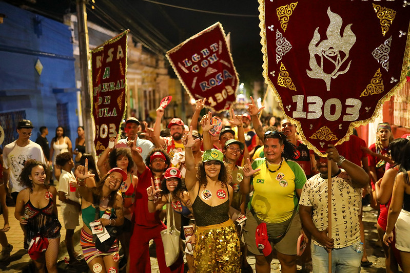 Foto noturna de uma caminhada ou manifestação em uma rua urbana, com um grupo de pessoas reunidas e posando para a câmera. Em primeiro plano, participantes vestem roupas coloridas, camisetas, bonés e adereços, enquanto várias pessoas levantam as mãos em gesto de saudação. Destacam-se grandes estandartes vermelhos com detalhes dourados e a inscrição “1303”, além de outros cartazes vermelhos carregados ao fundo. A cena é iluminada por luzes da rua e de estabelecimentos próximos, criando um ambiente movimentado e festivo, com muitas pessoas ocupando a via pública.
