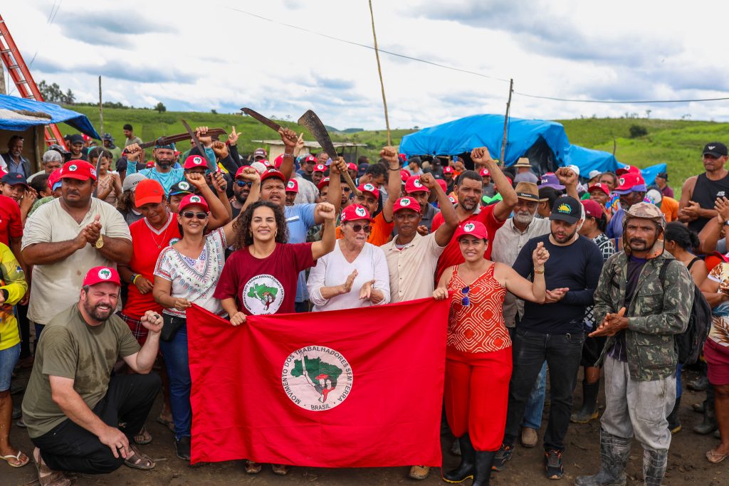 Foto de um grande grupo de pessoas reunidas em uma área rural ao ar livre, sob um céu nublado. Na frente, várias pessoas seguram uma bandeira vermelha com o símbolo do Movimento dos Trabalhadores Sem Terra (MST), enquanto outras levantam os braços ou aplaudem. Muitos participantes usam bonés vermelhos com o mesmo emblema, e algumas pessoas ao fundo seguram ferramentas agrícolas erguidas. Ao redor, há barracas cobertas por lonas azuis e uma paisagem de campos verdes, compondo uma cena de mobilização coletiva em ambiente de campo.