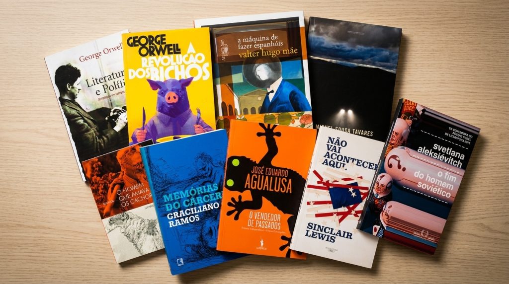 A imagem mostra uma seleção de livros dispostos sobre uma superfície de madeira clara, com capas coloridas e parcialmente sobrepostas. Entre os títulos visíveis estão obras de autores como George Orwell, Graciliano Ramos, José Eduardo Agualusa, Sinclair Lewis, Svetlana Aleksiévitch e Valter Hugo Mãe, reunindo diferentes gêneros e temas da literatura. As capas variam entre ilustrações, fotografias e designs gráficos marcantes, criando uma composição visual que destaca a diversidade e a riqueza do acervo apresentado.