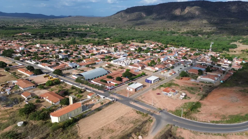 A imagem aérea mostra uma área plana com construções como casas e galpões. Ao fundo, uma serra coberta de vegetação de caatinga