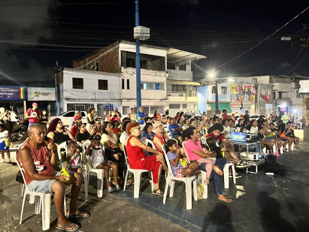 Foto noturna de uma atividade comunitária ao ar livre, com dezenas de pessoas de diferentes idades sentadas em cadeiras plásticas dispostas em uma praça ou rua, assistindo a uma projeção realizada por equipamentos montados em frente ao público. Ao fundo aparecem prédios residenciais e comerciais iluminados, postes de luz e pessoas em pé acompanhando a exibição, formando um ambiente de encontro cultural e convivência coletiva em espaço urbano.