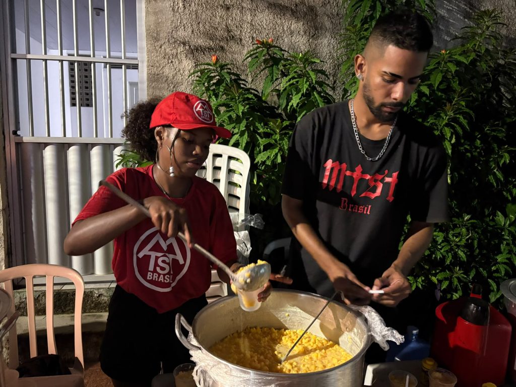 Foto de uma atividade comunitária noturna em que duas pessoas - uma mulher e um homem negros - servem alimento em copos plásticos a partir de uma grande panela colocada sobre uma mesa. Uma delas veste camiseta e boné vermelhos com a inscrição “MST Brasil”, enquanto a outra usa camiseta preta também com a mesma identificação; ao redor há cadeiras plásticas, recipientes para servir bebidas, plantas e a fachada de uma residência, compondo uma cena de organização e distribuição de alimentos em um evento coletivo.
