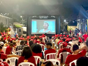 Foto de uma sessão de cinema ao ar livre realizada à noite em uma rua, com dezenas de pessoas sentadas em cadeiras plásticas voltadas para uma grande tela inflável com a inscrição “Cinema Sem Teto”. A maioria do público veste roupas e bonés vermelhos, enquanto a tela exibe a imagem de uma pessoa falando; ao redor, há postes de iluminação, construções urbanas e equipamentos de som, compondo um ambiente de evento comunitário e cultural.