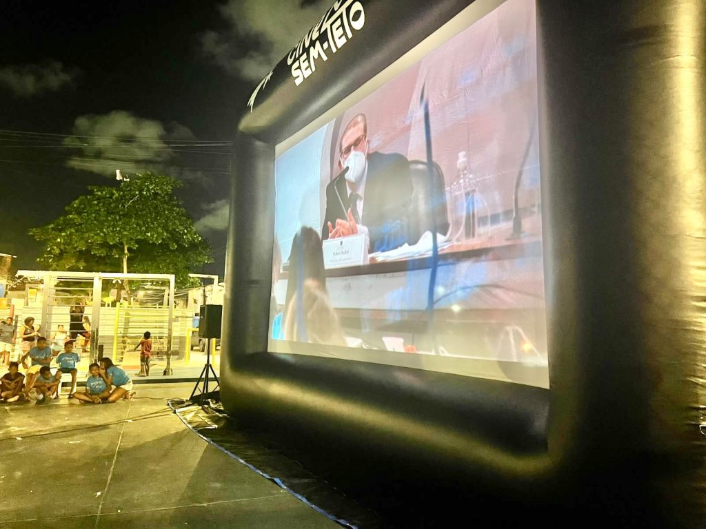 Foto noturna de uma exibição de cinema ao ar livre, com uma grande tela inflável identificada como “Cinema Sem Teto” ocupando a maior parte da cena. Na tela, aparece uma pessoa participando de uma reunião ou audiência, usando máscara e falando ao microfone, enquanto crianças e outras pessoas acompanham a projeção em um espaço público iluminado, cercado por árvores, postes de luz e estruturas urbanas ao fundo.