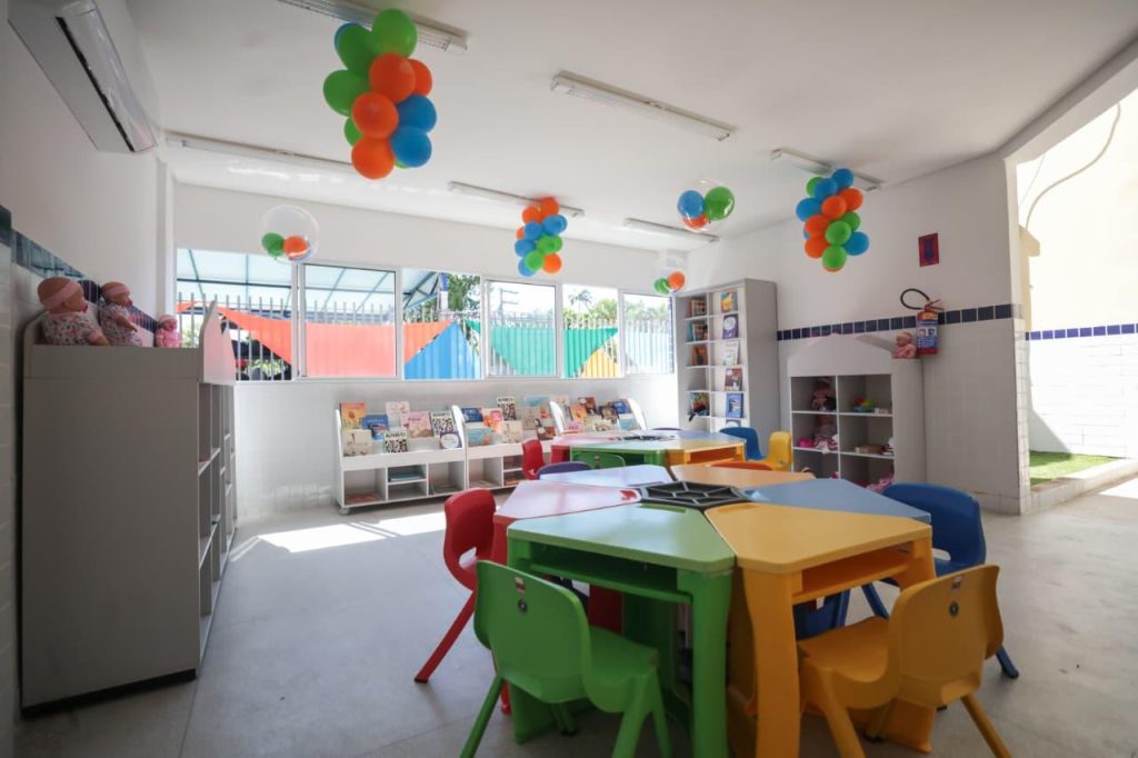 Sala infantil ampla, iluminada por luz natural, com mesas e cadeiras coloridas adequadas para crianças. O ambiente é decorado com balões nas cores verde, azul e laranja presos ao teto, e conta com estantes baixas contendo livros, brinquedos e bonecas. Ao fundo, grandes janelas permitem a entrada de luz e mostram uma área externa colorida. À direita, há um espaço de brincadeiras com nichos organizadores e acesso a uma área externa, criando um ambiente acolhedor e estimulante para atividades educativas e recreativas.