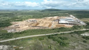 Vista aérea de uma ampla área em obras cercada por vegetação, com um grande edifício industrial em construção à direita, extensos trechos de solo terraplanado e equipamentos distribuídos pelo canteiro. Em primeiro plano, uma rodovia corta a paisagem, enquanto ao fundo se estendem áreas verdes e pequenas elevações sob um céu parcialmente nublado, destacando o contraste entre o desenvolvimento da infraestrutura e o ambiente natural ao redor.