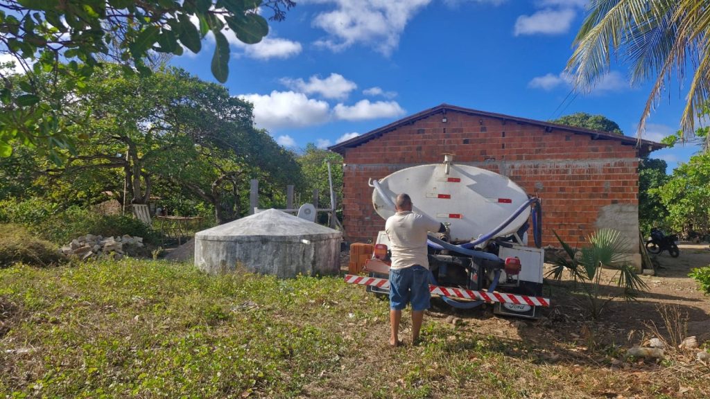 Foto de uma área rural com vegetação abundante e céu azul com nuvens brancas. Em primeiro plano, uma pessoa está posicionada atrás de um caminhão-tanque branco estacionado ao lado de uma construção simples de tijolos aparentes. Próximo ao veículo, há uma estrutura circular de concreto com cobertura em formato cônico, sugerindo um reservatório ou sistema de armazenamento de água. Árvores e coqueiros cercam o local, reforçando o ambiente campestre e de infraestrutura hídrica.