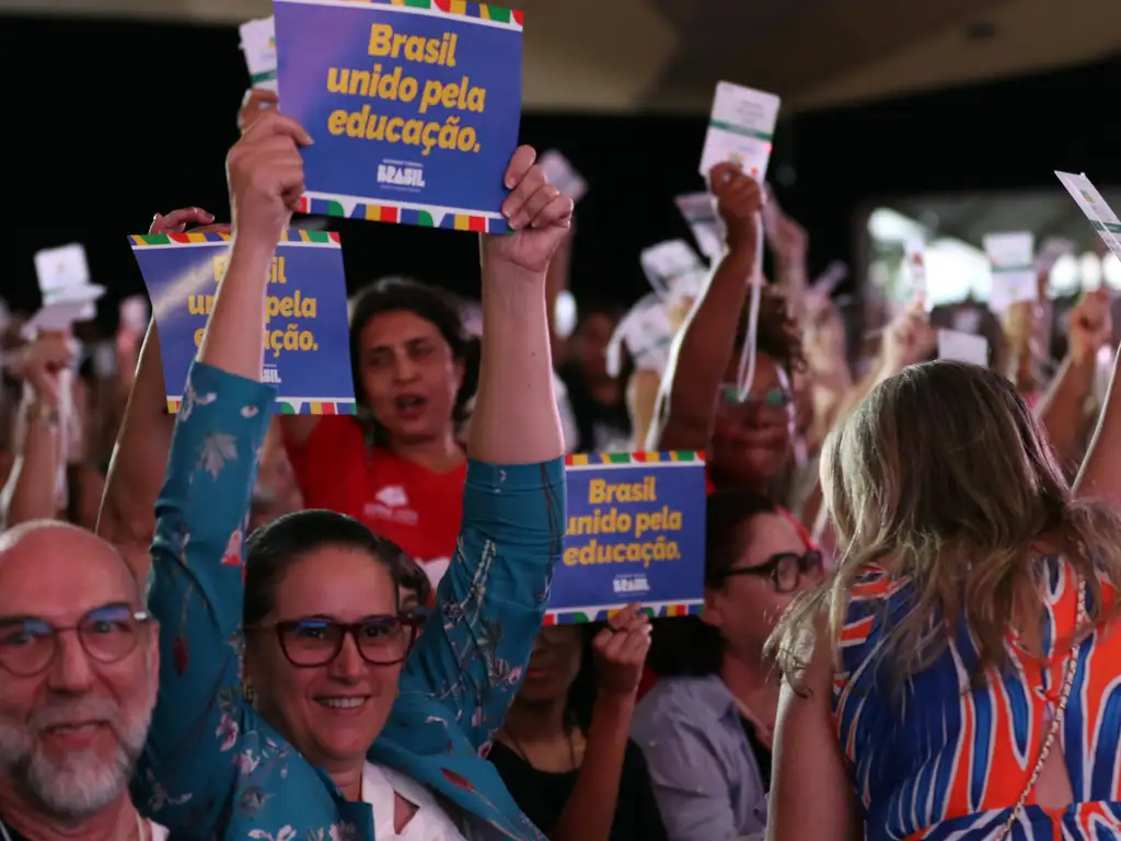 Em um evento com grande público, várias pessoas levantam placas azuis com a frase “Brasil unido pela educação”. Em primeiro plano, uma pessoa de óculos e roupa azul estampada ergue uma das placas acima da cabeça. Ao redor, outras pessoas também seguram placas e cartões, formando uma cena de mobilização coletiva em um ambiente iluminado e movimentado.