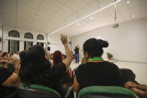Sala ampla e bem iluminada com várias pessoas sentadas em cadeiras, participando de uma atividade coletiva. Em primeiro plano, algumas pessoas aparecem de costas, e uma delas está com o braço levantado. Ao fundo, uma pessoa está em pé falando para o grupo, próxima a uma parede branca. O ambiente possui janelas altas, ventiladores nas paredes, iluminação de teto e um projetor suspenso. Há também uma planta em vaso em um canto da sala.
