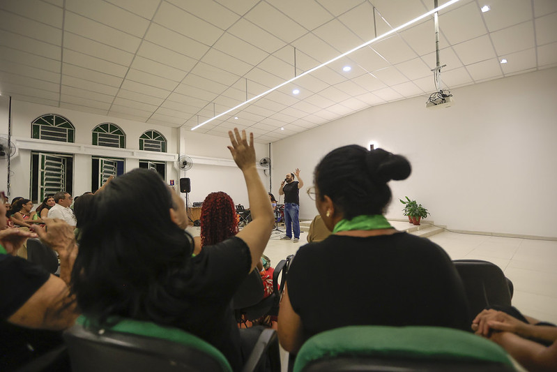 Sala ampla e bem iluminada com várias pessoas sentadas em cadeiras, participando de uma atividade coletiva. Em primeiro plano, algumas pessoas aparecem de costas, e uma delas está com o braço levantado. Ao fundo, uma pessoa está em pé falando para o grupo, próxima a uma parede branca. O ambiente possui janelas altas, ventiladores nas paredes, iluminação de teto e um projetor suspenso. Há também uma planta em vaso em um canto da sala.