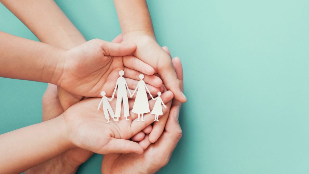 Mãos de diferentes tamanhos formam uma concha ao redor de um recorte branco de uma família de papel, composto por quatro figuras de mãos dadas, sobre um fundo azul-claro. A imagem simboliza proteção, cuidado, união e apoio familiar.
