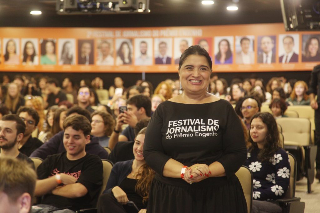A foto mostra uma mulher sorridente em pé diante de uma plateia em um evento de jornalismo, vestindo uma blusa preta com a identificação “Festival de Jornalismo – Prêmio Engenho”. Ao fundo, há diversas pessoas sentadas em um auditório, participando do evento.