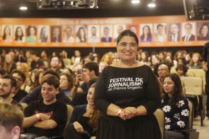 A foto mostra uma mulher sorridente em pé diante de uma plateia em um evento de jornalismo, vestindo uma blusa preta com a identificação “Festival de Jornalismo – Prêmio Engenho”. Ao fundo, há diversas pessoas sentadas em um auditório, participando do evento.
