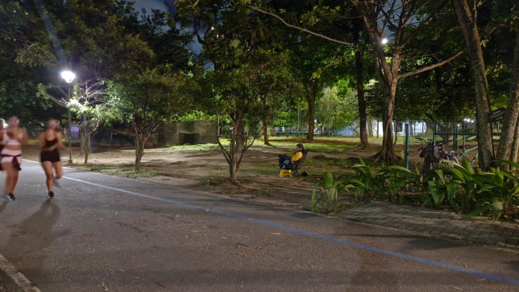 A imagem mostra, em plano aberto, o parque da Jaquiera à noite, com uma pessoa, um fotógrafo, sentado em uma cadeira voltado para a pista. duas pessoas corrrendo aparecem borradas na imagem