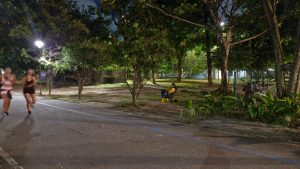 A imagem mostra, em plano aberto, o parque da Jaquiera à noite, com uma pessoa, um fotógrafo, sentado em uma cadeira voltado para a pista. duas pessoas corrrendo aparecem borradas na imagem