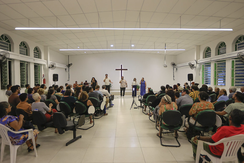 Vista ampla de um salão de igreja com fileiras de cadeiras ocupadas por pessoas voltadas para a frente; ao centro, um grupo conduz uma apresentação ou momento de louvor diante de uma cruz iluminada na parede, em um ambiente amplo, bem iluminado, com janelas laterais e atmosfera de reunião comunitária.
