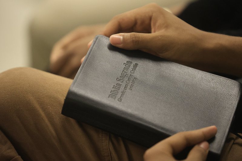 Close-up de duas mãos segurando uma Bíblia Sagrada de capa preta, apoiada sobre o colo de uma pessoa vestindo calça marrom; a imagem tem iluminação suave e fundo desfocado, destacando o livro e transmitindo um ambiente tranquilo e contemplativo.