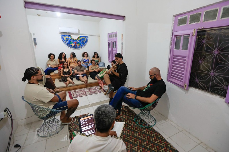 Vista de uma sala residencial simples com paredes brancas e janelas de moldura lilás, onde um grupo de pessoas está reunido em círculo e sentado em diferentes assentos, enquanto uma pessoa toca violão ao centro; o ambiente é iluminado, acolhedor e organizado para um encontro comunitário ou momento de conversa e reflexão.