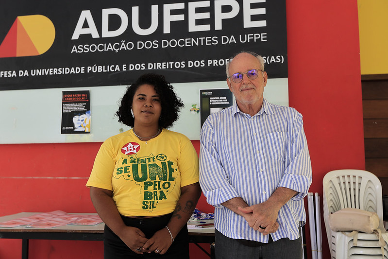 Duas pessoas estão lado a lado em um ambiente interno, em frente a um painel da ADUFEPE (Associação dos Docentes da UFPE). À esquerda, uma mulher jovem e negra usa camiseta amarela com texto em verde e vermelho. À direita, um homem branco idoso veste camisa listrada azul e branca e usa óculos. Ao fundo, há uma mesa com materiais impressos, cartazes na parede e cadeiras plásticas empilhadas. O espaço tem paredes em tons de vermelho, branco e amarelo.