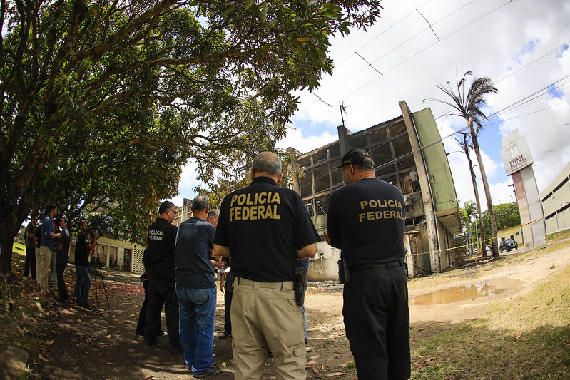 Em uma área externa arborizada, várias pessoas observam um prédio com sinais de danos causados por incêndio. Em primeiro plano, três pessoas vestem camisetas escuras com a inscrição “Polícia Federal” nas costas. Ao fundo, o edifício apresenta janelas sem vidro e marcas de queimadura na estrutura. Há árvores, uma área de terra com poças d’água e algumas pessoas registrando a cena com câmeras.