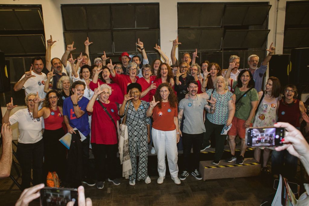 Grupo numeroso de pessoas posa para uma foto em ambiente interno, em frente a grandes janelas escuras. As pessoas estão reunidas em várias fileiras, muitas com um dos braços levantados fazendo um gesto em forma de “L” com a mão. As roupas são variadas e coloridas, com destaque para tons de vermelho, branco, azul e verde. No primeiro plano, aparecem mãos segurando celulares que registram a cena, enquanto o grupo ocupa o centro da imagem em uma composição coletiva de celebração e participação.