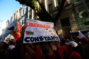 Uma manifestação ocorre em uma rua cercada por prédios antigos e árvores. Em primeiro plano, várias pessoas vestindo roupas vermelhas seguram um cartaz branco com a frase “LIBERDADE É UMA LUTA CONSTANTE” escrita em letras grandes. Também são visíveis bandeiras vermelhas entre a multidão. A cena acontece durante o dia, com áreas iluminadas pelo sol e outras em sombra, destacando o caráter coletivo e público do ato.