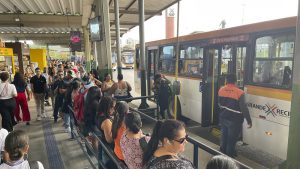 Foto de uma plataforma de terminal de ônibus movimentada, com muitas pessoas aguardando em fila ou próximas à área de embarque. À direita, um ônibus branco com faixas laranja está parado para o desembarque, enquanto uma pessoa uniformizada acompanha a operação. A estrutura do terminal é coberta por telhado metálico, com corrimãos organizando a circulação dos passageiros e sinalização visível ao longo da área. Ao fundo, é possível ver outro ônibus se aproximando, reforçando o intenso fluxo de transporte público no local.