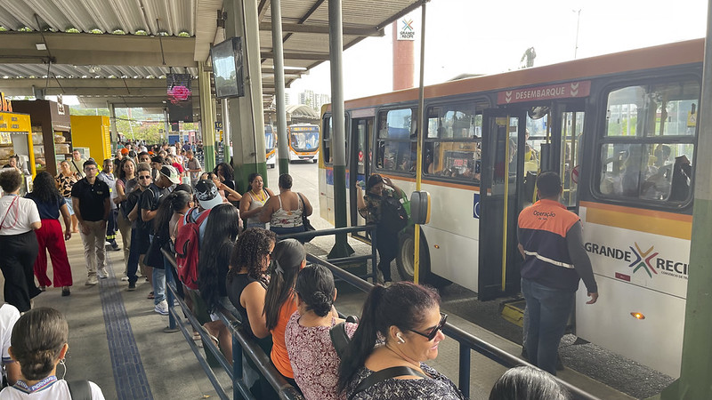 Foto de uma plataforma de terminal de ônibus movimentada, com muitas pessoas aguardando em fila ou próximas à área de embarque. À direita, um ônibus branco com faixas laranja está parado para o desembarque, enquanto uma pessoa uniformizada acompanha a operação. A estrutura do terminal é coberta por telhado metálico, com corrimãos organizando a circulação dos passageiros e sinalização visível ao longo da área. Ao fundo, é possível ver outro ônibus se aproximando, reforçando o intenso fluxo de transporte público no local.