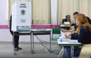 Em uma seção eleitoral, uma pessoa está usando uma cabine de votação branca identificada com os dizeres “Justiça Eleitoral” e “Cabina de Votação”. Ao lado, em mesas organizadas, três pessoas trabalham com documentos e equipamentos de apoio ao processo eleitoral. O ambiente é interno, com paredes claras, cortinas ao fundo e mobiliário simples, transmitindo o contexto de uma votação oficial.